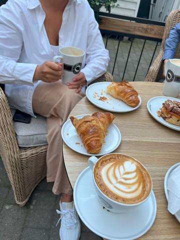 En kvinde i en hvid skjorte sidder på en cafe med en kop kaffe i hånden og en croissant på bordet, sammen med hendes nye veninde fra Boblberg